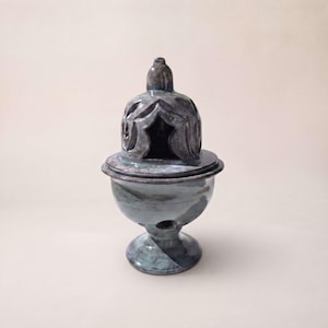 Op de afbeelding: Een keramische wierookbrander met een glanzend, gemarmerd blauw en grijs glazuur. Het object heeft een voetstuk, een afgerond komgedeelte met kleine ronde openingen en een afneembaar koepelvormig deksel met gedetailleerde boogvormige uitsparingen.