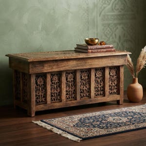 May include: A rectangular wooden storage chest featuring intricate floral carvings on the front panels and top lid. The chest has a natural brown finish and sits on four short legs. Two stacked books and a small gold bowl rest on the top surface.