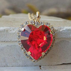 Puede incluir: Un colgante en forma de corazón con una gran piedra preciosa roja facetada engastada en un marco de metal dorado. El borde del colgante está rodeado por una hilera de pequeños cristales transparentes y brillantes. Una anilla dorada está unida a la parte superior.