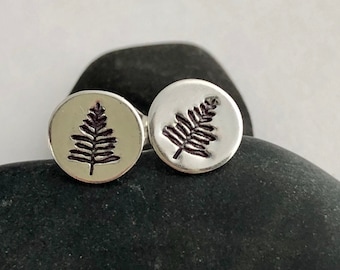 May include: A pair of silver-colored stud earrings. Each earring features a circular design with a detailed, dark brown fern or leaf imprint. The earrings are displayed on a dark, textured stone.