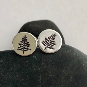 May include: A pair of silver-colored stud earrings. Each earring features a circular design with a detailed, dark brown fern or leaf imprint. The earrings are displayed on a dark, textured stone.
