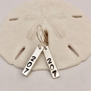 May include: A pair of silver rectangular earrings with the number "207" in black lettering. The earrings hang from silver hooks and are displayed on a white, textured surface.