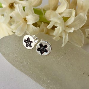 May include: A pair of silver stud earrings with a black starfish design. The earrings are on a white, textured surface.