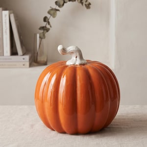 May include: A glossy, bright orange ceramic pumpkin decoration featuring deep vertical ridges and a smooth, curved white stem. The pumpkin has a rounded, bulbous shape and a reflective surface that catches the light, creating a vibrant autumn display.