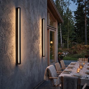 May include: Two vertical, rectangular black wall-mounted light fixtures with integrated LED strips that emit a warm, soft glow onto the textured gray exterior wall of a building, creating a linear illumination effect.
