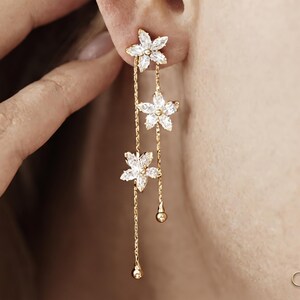 Puede incluir: Un pendiente colgante de tono dorado con tres dijes en forma de flor adornados con piedras transparentes de corte marquesa. El diseño incluye dos cadenas delicadas de diferentes longitudes, cada una terminada con una pequeña cuenta dorada. Texto GRACEGILD.