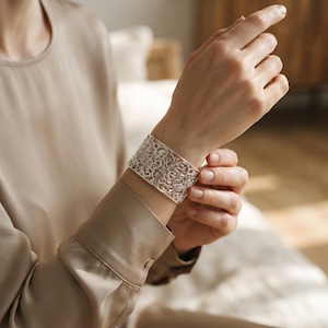 Puede incluir: Una pulsera de brazalete ancha de plata con un intrincado patrón de filigrana calada con bucles metálicos entrelazados. La superficie metálica pulida refleja la luz, resaltando el diseño recortado detallado en toda la banda.