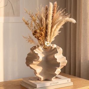 May include: A cream-colored ceramic vase with a wavy, shell-like rim containing a bouquet of dried grasses and flowers. The vase rests on two stacked books, with the top book clearly displaying the word AUTUMN on its white cover.