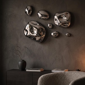 May include: A collection of six irregular, reflective silver wall sculptures with smooth, liquid-like surfaces. These metallic, pebble-shaped objects vary in size and are mounted in a cluster, creating a distorted mirror effect on the wall.