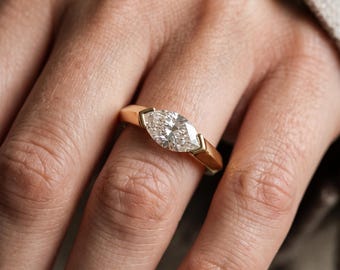 Può includere: Un anello in oro con una pietra preziosa trasparente taglio marquise, incastonata orizzontalmente in una montatura a tensione. La fascia è liscia e lucida, realizzata in oro giallo, offrendo un aspetto pulito che esalta la pietra.