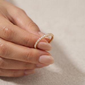 Puede incluir: Un anillo de banda fina de oro con una hilera continua de pequeñas piedras transparentes de corte redondo a lo largo de la superficie exterior. El metal tiene un acabado amarillo pulido y las piedras están engastadas en pavé, creando un aspecto uniforme.
