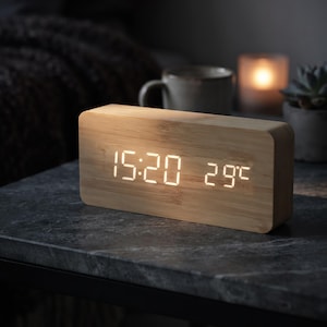 Puede incluir: Un reloj despertador digital rectangular con acabado en madera de bambú clara. La pantalla frontal presenta números LED de color blanco brillante que muestran la hora 15:20 y la temperatura de 29°C, integrados en la superficie de madera.
