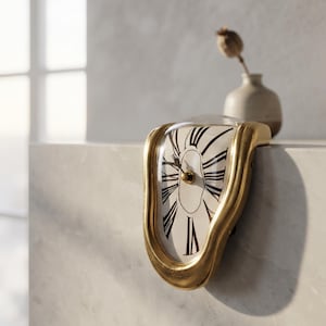 May include: A gold-toned melting-style clock with a white face, black Roman numerals, and black hands. The clock features a curved, distorted frame that drapes over the edge of a flat surface, creating a fluid, asymmetrical appearance.