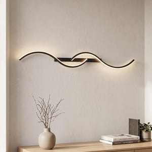 May include: A horizontal wall-mounted light fixture featuring two overlapping, curved black metal arms with integrated LED strips that emit a warm white glow, creating a continuous wave-like pattern across the wall surface.