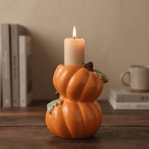 Può includere: Un portacandele in ceramica a forma di due zucche arancioni impilate con piccole foglie verdi. Una candela a colonna color crema è inserita nella zucca superiore, con una fiamma accesa che crea un bagliore caldo sulla superficie lucida.