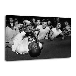 May include: A black and white photograph showing a musician lying on the floor playing a saxophone, surrounded by a crowd of people looking on. The musician wears a light-colored shirt, while the surrounding audience members watch intently.