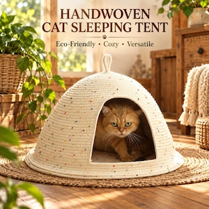 Pode incluir: Uma tenda de dormir para gatos em forma de cúpula, tecida à mão com corda de cor branco-sujo e pequenas manchas multicoloridas. A tenda apresenta uma abertura circular na frente e uma pequena alça no topo. Um gato ruivo está sentado no interior.