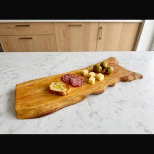 Può includere: Un tagliere rettangolare in legno con bordo naturale e venature a vista. Il tagliere presenta una fetta di pane tostato con olio, due fette di salame, quattro piccoli cubetti di formaggio giallo e tre olive verdi disposte sopra.