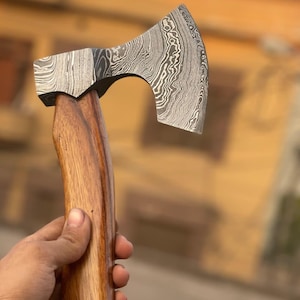 May include: A hand axe featuring a sharp, curved blade made of Damascus steel with a distinct wavy, layered pattern in shades of gray and black. The axe has a smooth, polished wooden handle with a natural brown grain and a curved ergonomic shape.