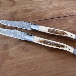 Pode incluir: Duas facas dobráveis com lâminas de aço Damasco apresentando um padrão ondulado e em camadas. Os cabos são feitos de osso de cor clara com uma incrustação central texturizada em castanho escuro e um reforço e tampa de metal prateado em cada faca.