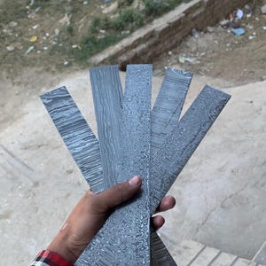 May include: Five rectangular metal billets featuring distinct Damascus steel patterns in shades of gray and black. Each piece displays a unique layered or swirling texture across its surface, arranged in a fan shape to showcase the metalwork.