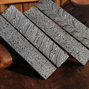 May include: Four rectangular metal bars featuring distinct Damascus steel patterns. Each bar displays a unique high-contrast black and silver layered design, ranging from intricate circular motifs to flowing, wavy lines across the flat surfaces.
