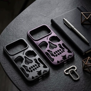 Op de afbeelding: Twee rechthoekige smartphonehoesjes met een uitgesneden schedelontwerp. De ene hoes is matzwart en de andere is metallic paars. Beide hoesjes hebben grote openingen voor cameralenzen en nauwkeurige uitsparingen voor de ogen, neus en tanden.