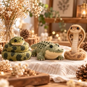 Può includere: Tre figure di animali in lana infeltrita: un serpente verde arrotolato con macchie scure, un alligatore verde con il dorso testurizzato e un cobra beige eretto con segni marroni. Ogni figura presenta piccoli occhi a perla neri e una consistenza morbida.