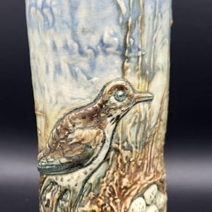 Puede incluir: Jarrón cilíndrico de cerámica con un relieve detallado de un pájaro y un nido. El pájaro es marrón y blanco, posado sobre un nido con huevos. El fondo presenta un cielo azul y troncos de árboles.