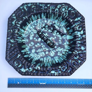 May include: A square ceramic ashtray with a dark brown and turquoise blue drip glaze pattern. The center features a raised black bar for resting items. A blue Victor ruler is positioned below, showing the ashtray is approximately 8 inches wide.