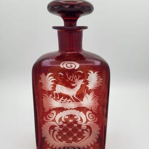 May include: A ruby red glass decanter with a stopper. The decanter features an etched design of a deer in a landscape with foliage and decorative scrollwork. The stopper is a rounded shape with a textured top.
