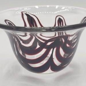May include: Clear glass bowl with a flared rim and a dark purple and white swirl design. The bowl's exterior features a pattern of curved lines, creating a decorative and artistic appearance. The bowl is suitable for decorative use.