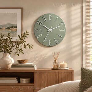 Puede incluir: Un reloj de pared redondo con una esfera de color verde salvia y números blancos en relieve. El reloj cuenta con manecillas blancas finas para las horas, los minutos y los segundos. El diseño es circular y muestra los números del uno al doce.