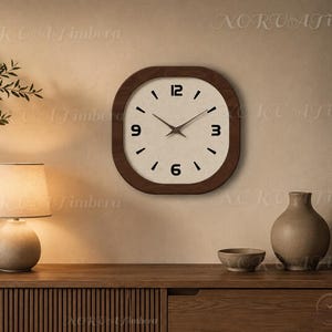 May include: A square wall clock with rounded corners, featuring a dark brown wooden frame and a light-coloured face. The clock has black numerals at the 12, 3, 6, and 9 positions, with simple black tick marks for the remaining hours and thin hands.