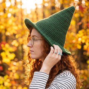 May include: A dark green crocheted pointed hat with a wide brim. The hat features a textured yarn construction with a tall, conical crown that tapers to a rounded point. The brim is slightly upturned at the front, showcasing the dense stitch pattern.