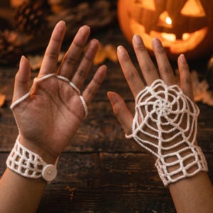 May include: A pair of white crochet fingerless gloves featuring a spider web pattern on the back of the hand. The gloves include a wrist cuff with a single white button closure and loops that secure around the fingers to hold the web design in place.