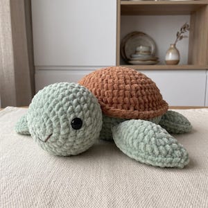 Op de afbeelding: Een pluche schildpadspeeltje gemaakt van zacht, grof garen. De schildpad heeft een ronde, licht saliegroene kop met één zichtbaar zwart plastic oog en een gestikte mond, gecombineerd met een textuurrijk, rond schild in een aardse bruine tint.