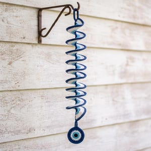 May include: A spiral-shaped crochet hanging decoration featuring a repeating pattern of dark blue, light blue, and white concentric circles. The structure ends with a circular evil eye charm at the bottom, suspended from a metal wall bracket.