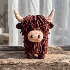 Op de afbeelding: Een handgemaakte gehaakte Highland-koe knuffel met lange, ruige donkerbruine garenvacht, een lichtbeige snuit, twee gebogen lichtbeige hoorns en kleine zwarte veiligheidsoogjes. Het knuffeldier heeft een dichte, getextureerde uitstraling.