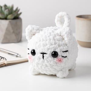 Puede incluir: Un peluche de gato tejido a ganchillo en color blanco con cuerpo redondeado, orejas pequeñas y cola enroscada. El gato tiene grandes ojos de plástico negro, nariz y boca bordadas en negro y mejillas rosadas. Textura suave y de lana.