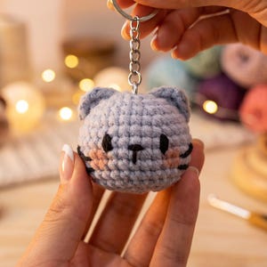May include: A light blue crocheted cat head keyring featuring black embroidered eyes, a small black nose, and pink stitched cheeks. The round, textured yarn figure is attached to a silver-toned metal chain and a circular split ring for keys.