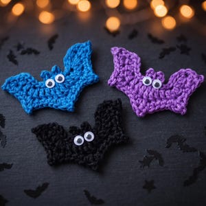 Può includere: Tre pipistrelli all'uncinetto in blu, viola e nero. Ogni pipistrello presenta un corpo in filato testurizzato con due occhi mobili bianchi con pupille nere fissati al centro. I pipistrelli sono disposti in gruppo, mostrando ali e orecchie.