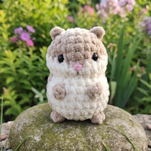 May include: A small, round, crocheted plush toy shaped like a hamster. The creature features a soft, textured beige and cream-colored yarn body, two small rounded ears, tiny front paws, and a small pink stitched nose with two black bead eyes.