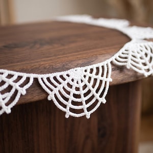 May include: A white crocheted garland featuring a repeating pattern of semi-circular spider web motifs. The delicate yarn construction creates a series of interconnected, fan-shaped segments that drape along the edge of a dark wooden surface.