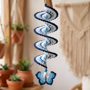 May include: A hanging spiral wind spinner crafted from crochet yarn in shades of navy blue, light blue, and white. The spiral structure features a scalloped edge and terminates in a blue and white butterfly motif with a small blue bead accent.