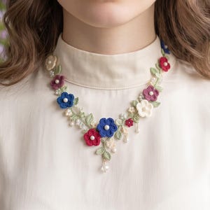 Puede incluir: Un collar floral de ganchillo con pequeñas flores rojas, azules, blancas y moradas intercaladas con hojas verdes de ganchillo y pequeñas perlas blancas. El collar tiene forma de V y descansa sobre una camisa de cuello alto color crema.