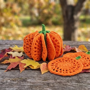 May include: A three-dimensional crocheted orange pumpkin with a green stem, accompanied by two flat, circular orange crocheted coasters featuring a textured, open-work stitch pattern, all arranged among scattered, colorful autumn leaves.