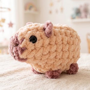 Peut inclure: Une peluche au crochet en forme de capybara, fabriquée à partir de fil beige clair doux avec des accents mauve foncé sur le museau, les oreilles et les pattes. Le jouet présente un corps arrondi, de petites oreilles et un œil en plastique noir.