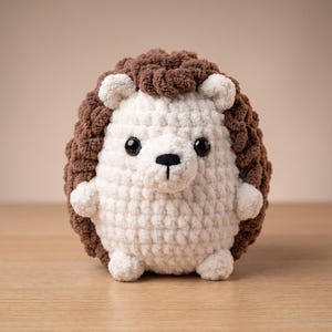 May include: A crochet hedgehog plush toy featuring a cream-coloured face, belly, and limbs with a textured dark brown back. The stuffed animal has two small black plastic eyes and a stitched black nose and mouth on its rounded face.