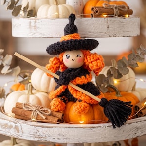 May include: A small crocheted witch doll with orange hair and a black hat, wearing a black and orange dress. The doll has a smooth wooden face with a winking expression and holds a miniature broomstick made of a wooden handle and black yarn.